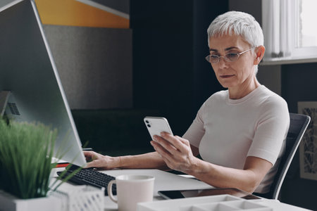 Confident senior businesswoman using smart phone while sitting at her working place in officeの写真素材