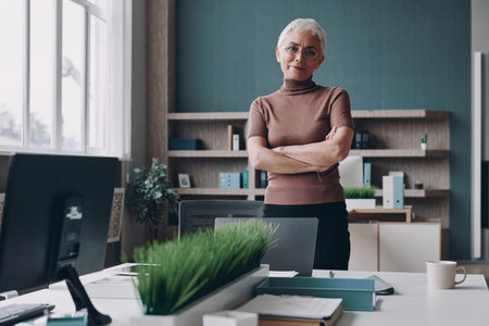 Senior businesswoman keeping arms crossed while standing near her working place in office