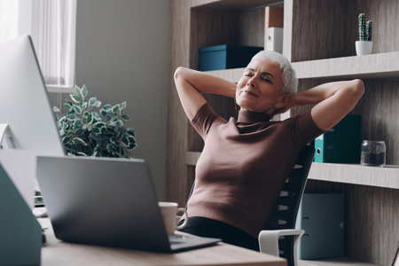 Relaxed senior businesswoman holding hands behind head while sitting at her working placeの写真素材
