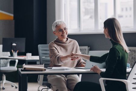 Two confident women shaking hands and smiling while sitting at the office desk togetherの写真素材