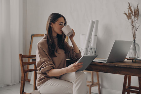 Young woman drinking coffee and looking at digital tablet while working in creative officeの写真素材