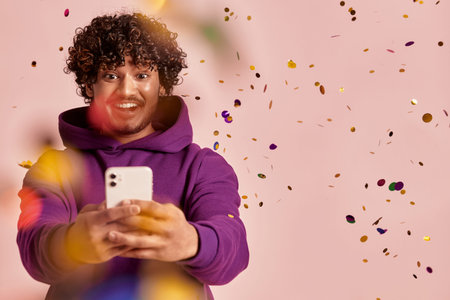 Joyful Indian man using smart phone while confetti flying around against pink background