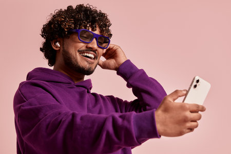 Happy Indian man adjusting headphones while using smart phone against pink backgroundの写真素材