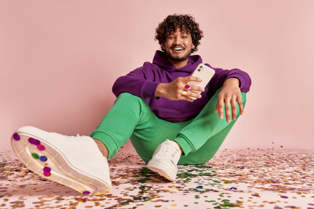 Happy Indian man holding smart phone while sitting on confetti against pink background
