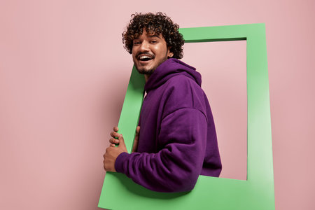 Happy Indian man looking out of the picture frame while standing against pink background