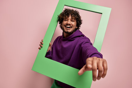 Happy Indian man looking out of the picture frame and pointing camera against pink backgroundの写真素材