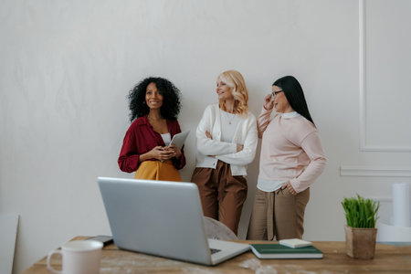 Three mature women communicating and smiling while leaning on the wall in officeの写真素材