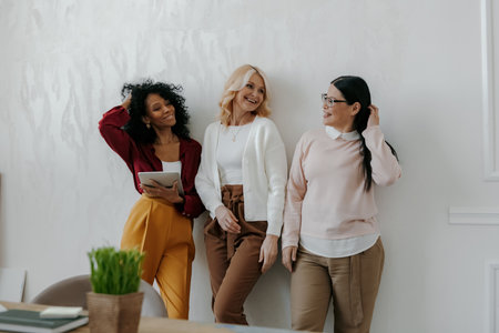 Three confident mature women talking and smiling while leaning on the wall in officeの写真素材