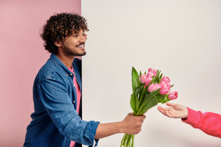 Happy Indian man giving bouquet of tulips to some woman against pink backgroundの写真素材