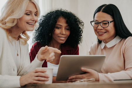 Three happy mature women looking at digital tablet while sitting at the desk togetherの写真素材