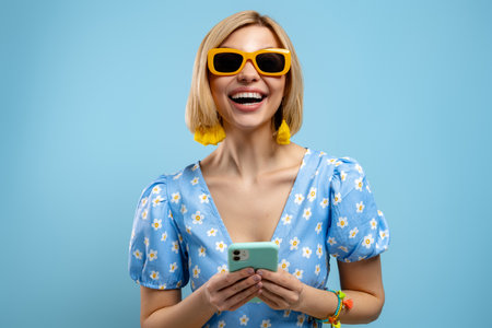 Excited young woman holding smart phone and smiling against blue backgroundの写真素材