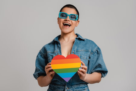 Happy young short hair woman holding heart shaped rainbow flag against grey backgroundの写真素材