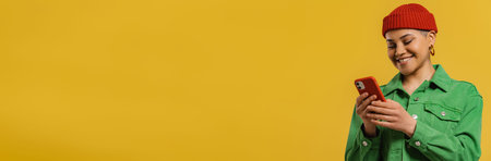Fashionable young woman using smart phone and smiling while standing against yellow background