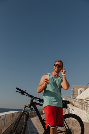 Happy young man in headphones leaning on his bicycle and smiling while spending time seasideの写真素材