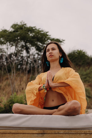 Confident young woman in boho styled crop top sitting in lotus position while meditating seasideの写真素材