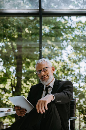Confident mature businessman holding digital tablet and smiling while sitting in the officeの写真素材