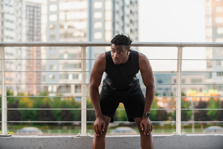 Tired African man in sportswear and headphones resting after sport training outdoors