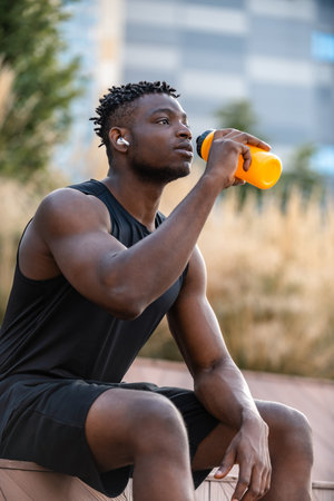 Handsome African man in sportswear drinking water while resting after training outdoors