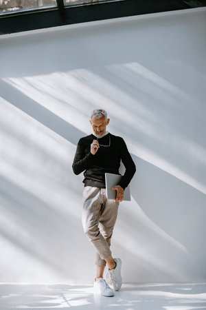 Full length of senior gray hair man in smart casual wear holding laptop while standing against a...