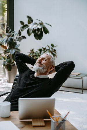 Happy mature businessman holding hands behind head and keeping eyes closed while resting in officeの写真素材