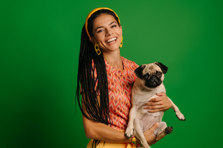 Joyful young Hispanic woman carrying cute pug dog and smiling against green backgroundの写真素材