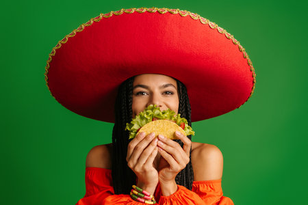 Cheerful Mexican woman in Sombrero hiding half face behind taco against green backgroundの写真素材