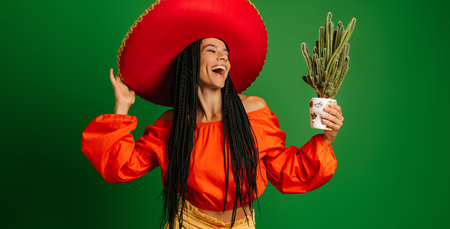 Beautiful Mexican woman in Sombrero holding cactus and looking at it with smile against green backgroundの写真素材
