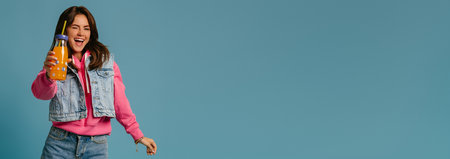 Happy young woman in denim wear holding bottle with lemonade and winking against blue backgroundの写真素材