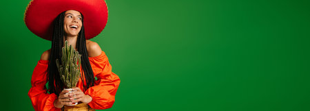 Joyful young Mexican woman in Sombrero holding cactus against green background