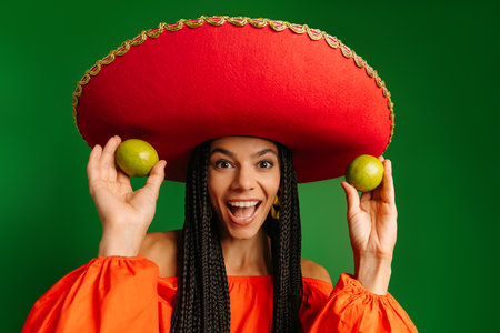 Joyful young Mexican woman in Sombrero showing fresh limes and smiling against green backgroundの写真素材