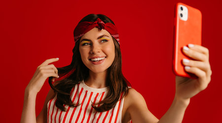 Beautiful young hipster woman making selfie by mart phone and smiling while standing on red backgroundの写真素材