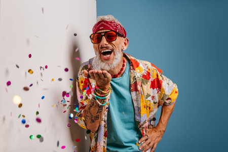 Happy senior man in funky shirt and bandana blowing colorful confetti on white and blue background