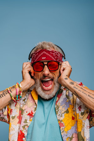 Hipster senior man in adjusting headphones and shouting while listening to the music against blue...