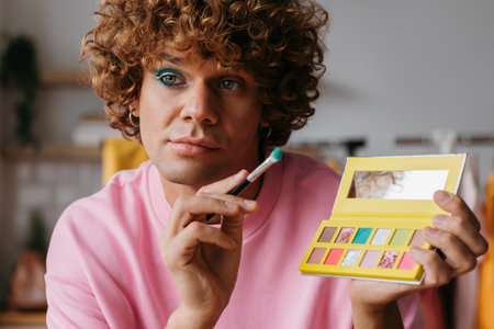 Confident young gay man holding eyeshadow palette while presenting make-up products from homeの写真素材