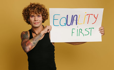 Handsome young gay man with make-up showing colorful banner against yellow background