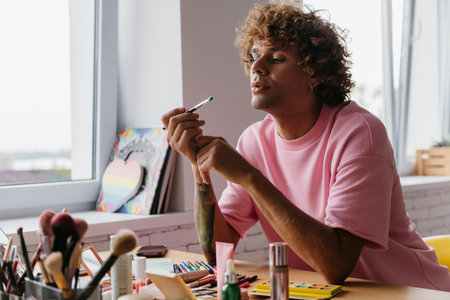 Confident young gay man blowing on eyeshadow brush while applying make-up at homeの写真素材
