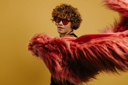 Curly young gay man in trendy glasses playing with fluffy coat against yellow backgroundの写真素材