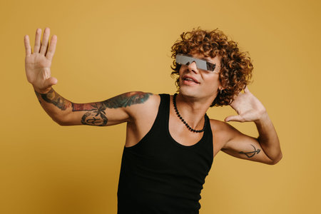 Fashionable curly young man in trendy glasses gesturing and smiling against yellow background