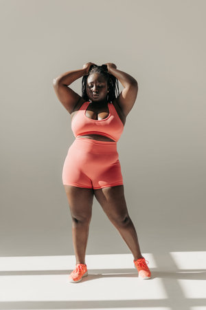 Full length of confident African plus size woman in sportswear adjusting her dreadlocs while...