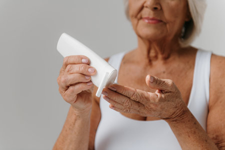 Close-up of senior woman pouring cosmetic cream on fingers against gray backgroundの写真素材