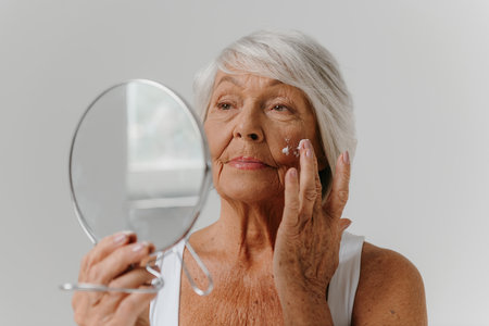 Confident senior woman using mirror while applying anti-age cream on face against gray backgroundの写真素材