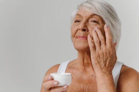 Confident senior woman applying anti-aging cream on face against gray backgroundの写真素材