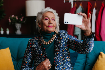 Fashionable senior woman photographing herself by smart phone while relaxing at modern living roomの写真素材