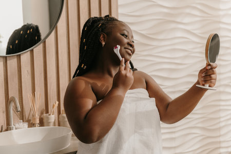 Confident plus size African woman covered in towel massaging face with roller at the domestic bathroomの写真素材
