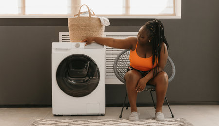 Young African woman select settings for laundry while sitting in comfortable chair near the washing machineの写真素材