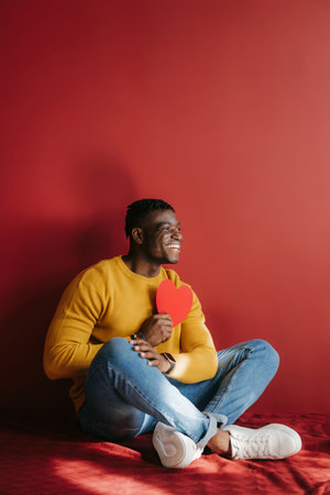 Happy young African man holding heart shaped Valentine card while sitting on the floor againstの写真素材