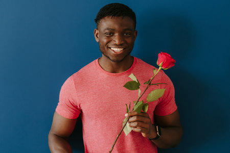 Handsome young African man in casual wear holding single rose and smiling on blue backgroundの写真素材
