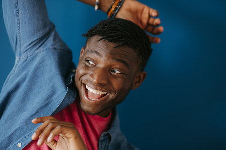Playful young African man in casual wear smiling and gesturing on blue backgroundの写真素材