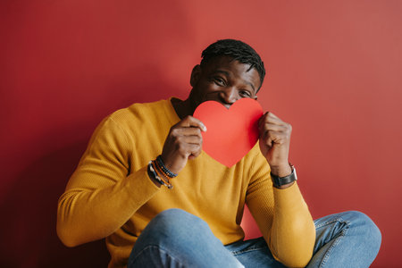 Playful young African man holding heart shaped Valentine card near face while sitting againstの写真素材