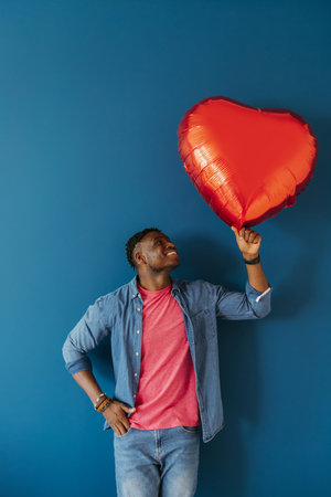 Handsome young African man holding heart shaped balloon and smiling while standing on red backgroundの写真素材